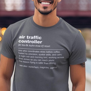 Könnte beinhalten: Ein dunkelgraues T-Shirt mit einem weißen Grafikdesign, auf dem "Fluglotse" steht und der Begriff definiert wird. Das Shirt wird von einer Person getragen, die auf einem Holzsteg steht.