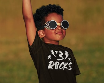 May include: A young person wearing goggles and a black t-shirt with the text "⚡️⚡️⚡️ ROCKS" holds a toy airplane above their head.