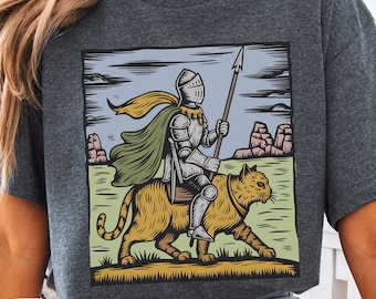 May include: Dark heather gray t-shirt featuring a graphic of a knight riding a tiger, holding a spear. The illustration has a medieval theme with a green cape and helmet. The text "DARK HEATHER" is displayed on the bottom.