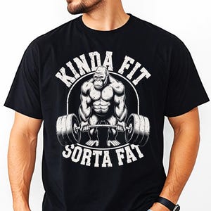 May include: Black t-shirt with a graphic of a muscular gorilla lifting a barbell. The text "KINDA FIT" arches above the gorilla, and "SORTA FAT" is below. The design is in white, creating a bold contrast against the black fabric.