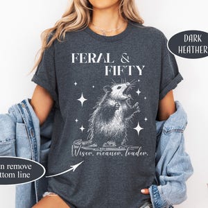 May include: Dark heather gray t-shirt with the text "FERAL & FIFTY" and a graphic of an opossum. The opossum is surrounded by stars and the text "Wiser, meaner, louder." is below the opossum.