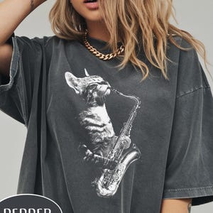 May include: Dark gray t-shirt featuring a black and white illustration of a cat playing a saxophone. The word "PEPPER" is printed in a white oval on the bottom of the shirt. The shirt has short sleeves and a relaxed fit.