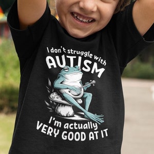 May include: A black t-shirt with a white graphic of a frog playing a banjo. The text on the shirt reads "I don't struggle with autism, I'm actually very good at it."