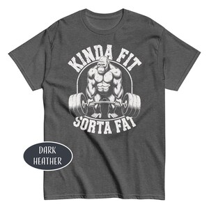 May include: Dark heather gray t-shirt with a white graphic of a gorilla lifting a barbell, with the text "KINDA FIT" above and "SORTA FAT" below. The shirt is a crew neck style.