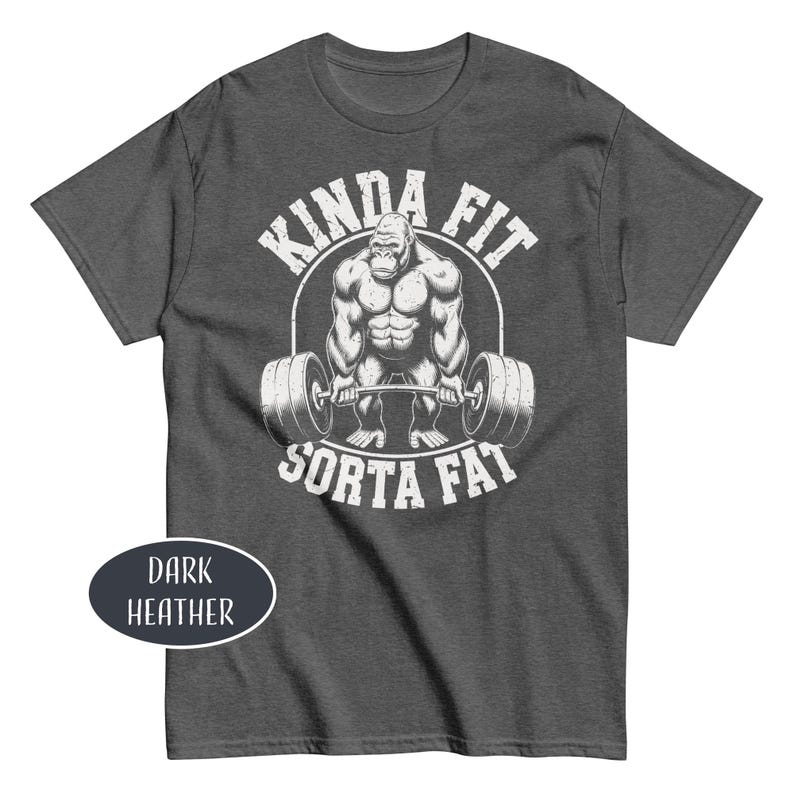 May include: Dark heather gray t-shirt with a white graphic of a gorilla lifting a barbell, with the text "KINDA FIT" above and "SORTA FAT" below. The shirt is a crew neck style.
