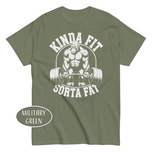 May include: Military green t-shirt featuring a muscular gorilla lifting a barbell. The graphic includes the text "KINDA FIT" above the gorilla and "SORTA FAT" below. The shirt's color is labeled "MILITARY GREEN".