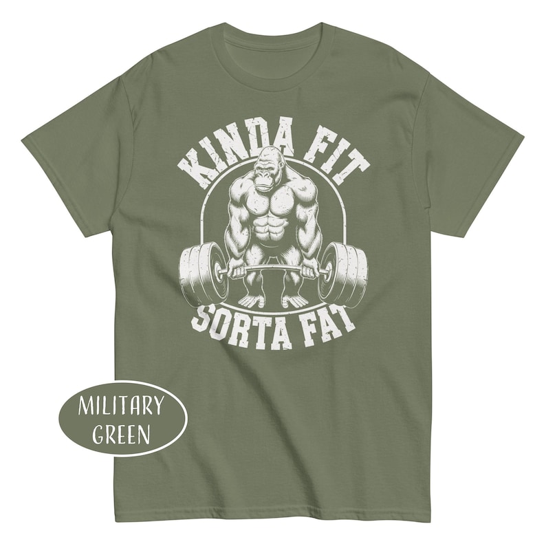 May include: Military green t-shirt featuring a muscular gorilla lifting a barbell. The graphic includes the text "KINDA FIT" above the gorilla and "SORTA FAT" below. The shirt's color is labeled "MILITARY GREEN".