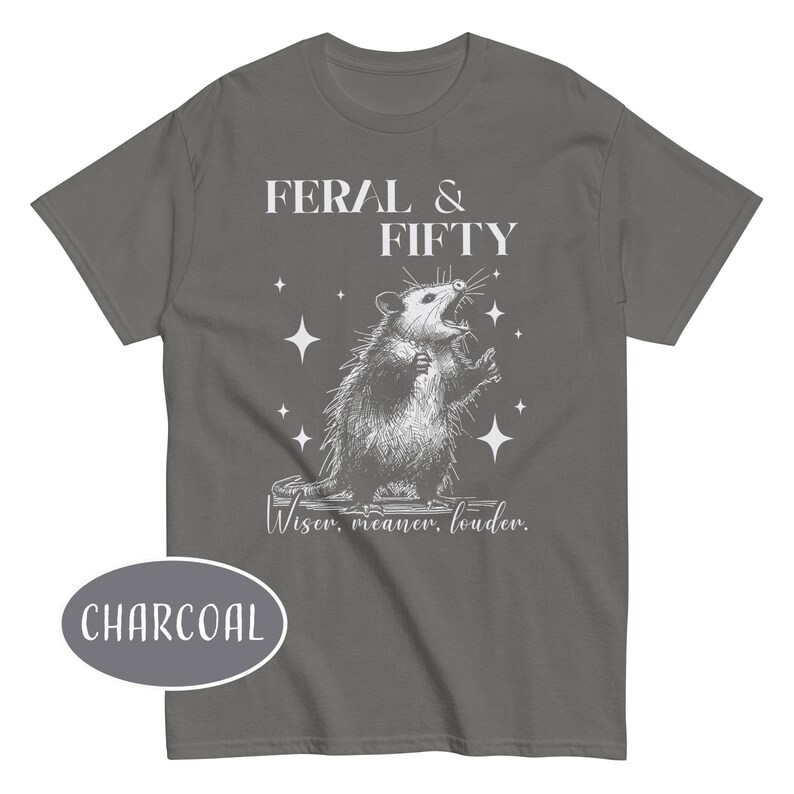 May include: Charcoal gray t-shirt with a graphic of an opossum with the text "FERAL & FIFTY" above it. Below the opossum is the text "Wiser, meaner, louder."