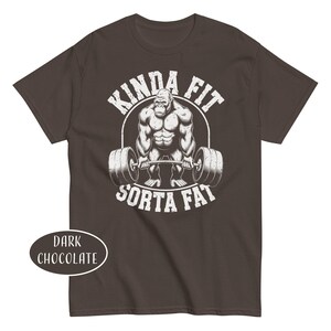 May include: Dark chocolate-colored t-shirt with a graphic of a weightlifting gorilla inside a white circle. The text "KINDA FIT" arches above the gorilla, and "SORTA FAT" is below. The shirt color is labeled "DARK CHOCOLATE".