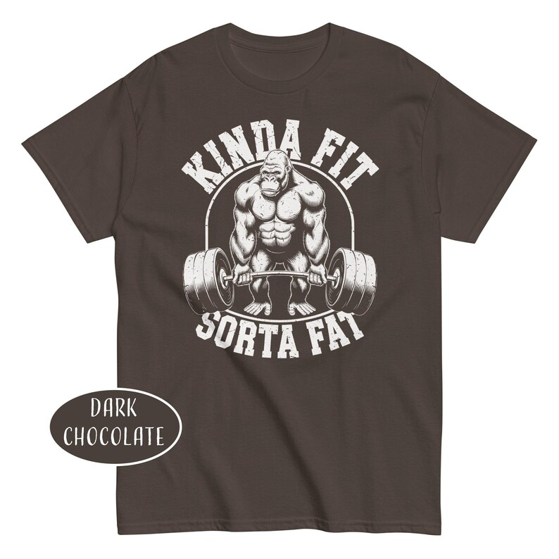 May include: Dark chocolate-colored t-shirt with a graphic of a weightlifting gorilla inside a white circle. The text "KINDA FIT" arches above the gorilla, and "SORTA FAT" is below. The shirt color is labeled "DARK CHOCOLATE".
