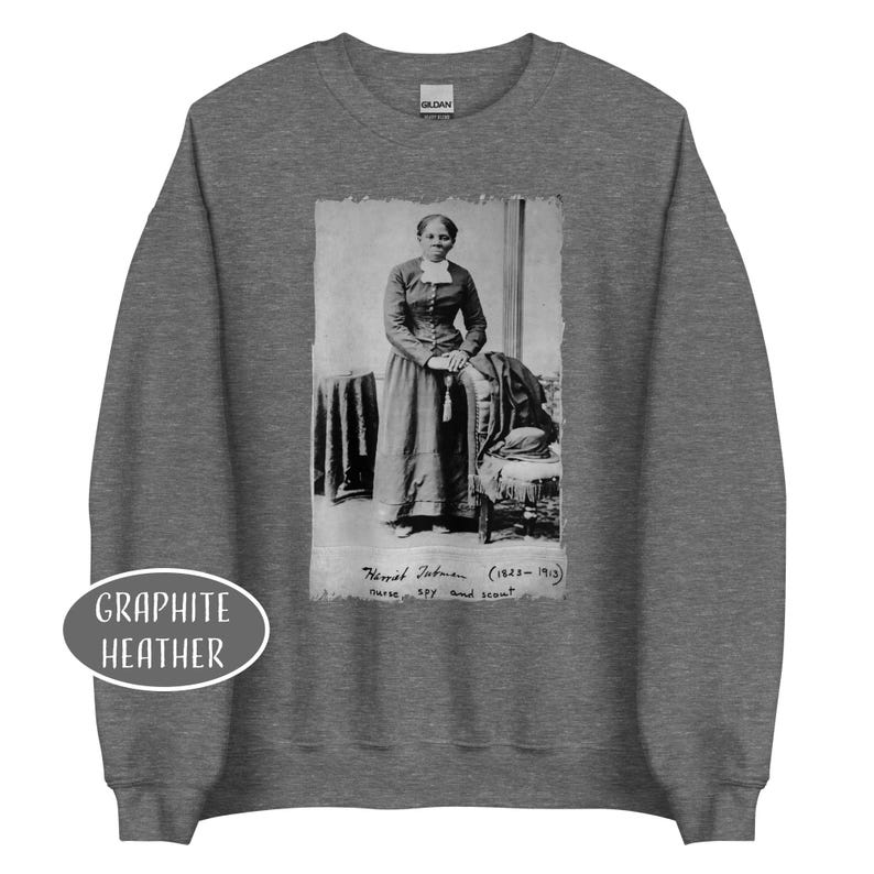 May include: Graphite heather sweatshirt featuring a black and white image of Harriet Tubman. The image includes text that reads "Harriet Tubman (1822-1913) nurse, spy and scout". The sweatshirt has a crew neck and long sleeves.