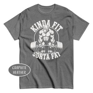 May include: Graphite heather t-shirt with a graphic of a gorilla lifting weights, encircled by the words "KINDA FIT" above and "SORTA FAT" below. The shirt is a dark gray color.
