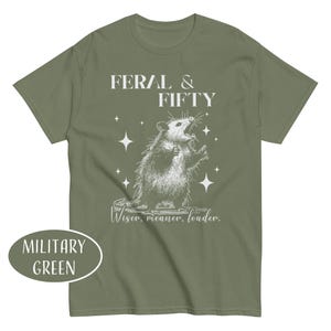 May include: Military green t-shirt with a graphic of an opossum and the text "FERAL & FIFTY". The opossum is drawn in a detailed, vintage style, with the words "Wiser, meaner, louder." below the graphic. The shirt is a solid color.