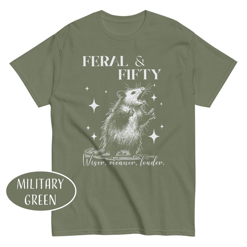 May include: Military green t-shirt with a graphic of an opossum and the text "FERAL & FIFTY". The opossum is drawn in a detailed, vintage style, with the words "Wiser, meaner, louder." below the graphic. The shirt is a solid color.
