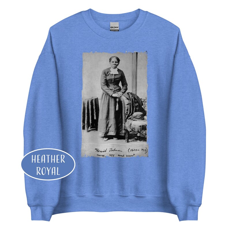May include: Heather royal blue sweatshirt featuring a black and white image of Harriet Tubman. The image includes text that reads "Harriet Tubman (1822-1913) nurse, spy, and scout." A white oval with the words "Heather Royal" is on the bottom left.