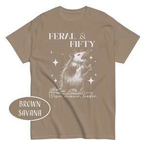 May include: Brown Savana t-shirt with a graphic of an opossum and the text "FERAL & FIFTY". The opossum is drawn in detail, with its mouth open and surrounded by star shapes. The text "Wiser, meaner, louder" is below the opossum.