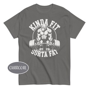 May include: Charcoal gray t-shirt featuring a muscular gorilla lifting a barbell. The text "KINDA FIT SORTA FAT" is printed above and below the gorilla in a distressed white font. A charcoal oval with the word "CHARCOAL" is in the lower left corner.