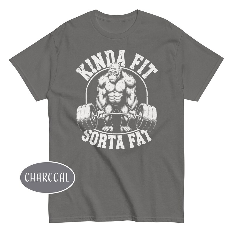 May include: Charcoal gray t-shirt featuring a muscular gorilla lifting a barbell. The text "KINDA FIT SORTA FAT" is printed above and below the gorilla in a distressed white font. A charcoal oval with the word "CHARCOAL" is in the lower left corner.