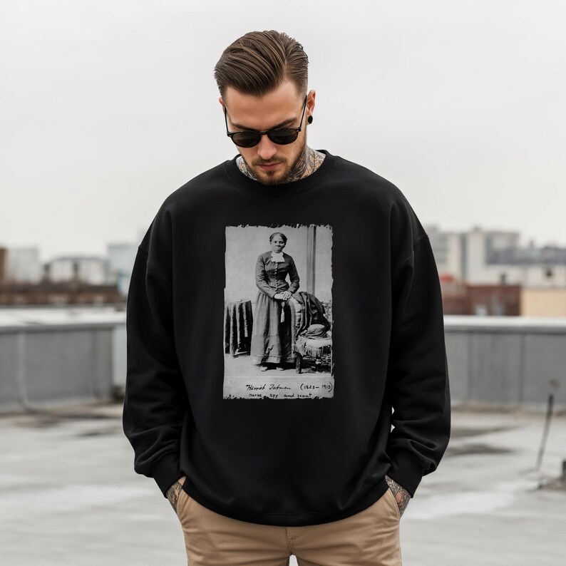 May include: Black sweatshirt featuring a black and white image of Harriet Tubman. The image is a portrait of Tubman standing next to a chair. The sweatshirt is worn by a person wearing sunglasses.