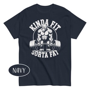 May include: Navy blue t-shirt with a white graphic of a gorilla lifting weights. The text "KINDA FIT" arches above the gorilla, and "SORTA FAT" is below. The shirt is a short-sleeved crew neck.