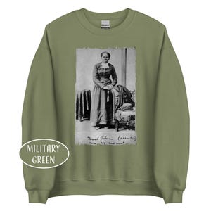 May include: Military green sweatshirt featuring a black and white portrait of Harriet Tubman. The portrait includes the text "Harriet Tubman (1823-1913) nurse, spy and scout". The sweatshirt has a crew neck and long sleeves.