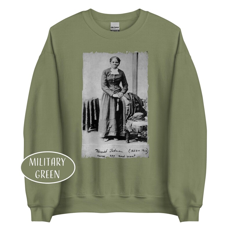 May include: Military green sweatshirt featuring a black and white portrait of Harriet Tubman. The portrait includes the text "Harriet Tubman (1823-1913) nurse, spy and scout". The sweatshirt has a crew neck and long sleeves.