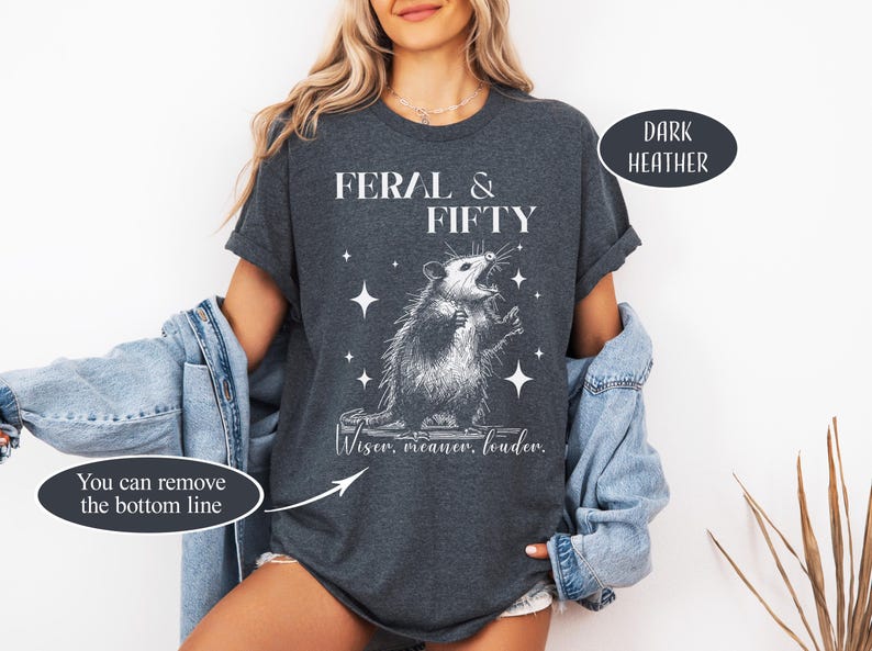 May include: Dark heather gray t-shirt with the text "FERAL & FIFTY" and a graphic of an opossum. The opossum is surrounded by stars and the text "Wiser, meaner, louder." is below the opossum.