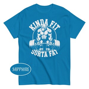 May include: Sapphire blue t-shirt with a white graphic of a gorilla lifting a barbell. The text "KINDA FIT" arches above the gorilla, and "SORTA FAT" is below. The shirt is a short-sleeved crew neck.