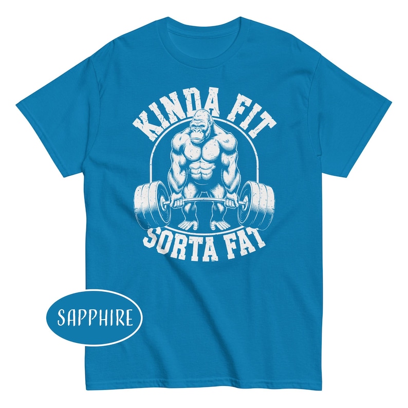 May include: Sapphire blue t-shirt with a white graphic of a gorilla lifting a barbell. The text "KINDA FIT" arches above the gorilla, and "SORTA FAT" is below. The shirt is a short-sleeved crew neck.