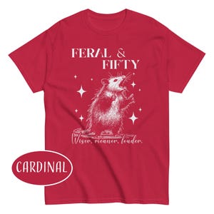 May include: Cardinal red t-shirt with a white graphic of an opossum and the text "FERAL & FIFTY". Below the graphic is the text "Wiser, meaner, louder."