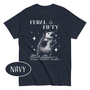 May include: Navy blue t-shirt with a graphic of an opossum and the text "FERAL & FIFTY". The opossum is illustrated in black and white, with the words "Wiser, meaner, louder" below the graphic. The word "NAVY" is in a white oval.