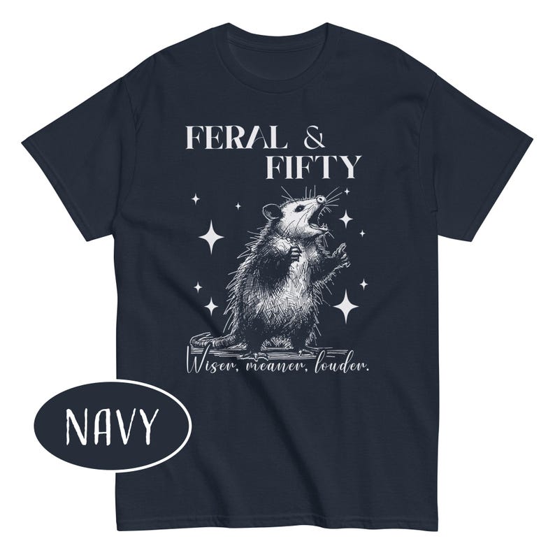 May include: Navy blue t-shirt with a graphic of an opossum and the text "FERAL & FIFTY". The opossum is illustrated in black and white, with the words "Wiser, meaner, louder" below the graphic. The word "NAVY" is in a white oval.