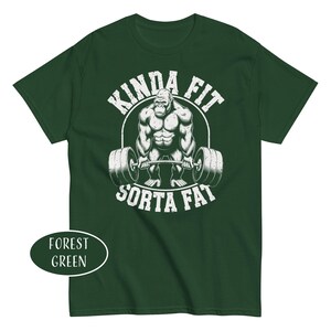 May include: Forest green t-shirt featuring a muscular gorilla lifting a barbell. The text "KINDA FIT" arches above the gorilla, with "SORTA FAT" below. The shirt's color is indicated by a label in the lower left corner.