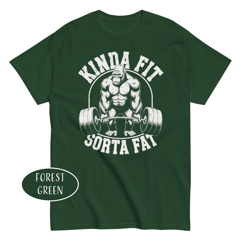 May include: Forest green t-shirt featuring a muscular gorilla lifting a barbell. The text "KINDA FIT" arches above the gorilla, with "SORTA FAT" below. The shirt's color is indicated by a label in the lower left corner.