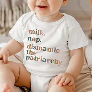 May include: White baby onesie with the text "milk. nap. dismantle the patriarchy." in various colors. The onesie is short-sleeved and has a simple, classic design. The text is in a playful, yet bold font.