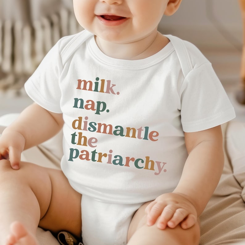 May include: White baby onesie with the text "milk. nap. dismantle the patriarchy." in various colors. The onesie is short-sleeved and has a simple, classic design. The text is in a playful, yet bold font.