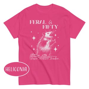 May include: A bright pink t-shirt featuring a white graphic of an opossum with the text "FERAL & FIFTY" above it. Below the opossum is the text "Wiser, meaner, louder." The shirt also has the word "HELICONIA" in a pink oval.