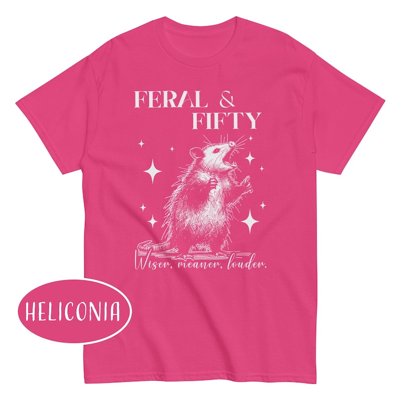 May include: A bright pink t-shirt featuring a white graphic of an opossum with the text "FERAL & FIFTY" above it. Below the opossum is the text "Wiser, meaner, louder." The shirt also has the word "HELICONIA" in a pink oval.