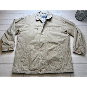 May include: Tan button-up jacket with a collared neckline and buttoned cuffs. The jacket has two front pockets and a plaid lining visible at the collar. The buttons are a matching tan color. The jacket is laid flat on a light wood surface.