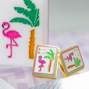 May include: A white Mahjong tile with a pink flamingo, green palm tree, and the number 1. Two gold-rimmed square earrings with a flamingo and palm tree design. The earrings have the text "Bird Bam".