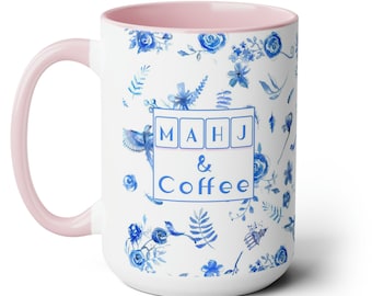 May include: White ceramic coffee mug with a pink interior and handle. The mug features blue floral patterns and the words "MAHJ & Coffee" in a square design. The design includes blue flowers, leaves, and birds.