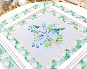 May include: A Christmas-themed table setting featuring a square print with a blue bow and holly design, framed by white wooden pieces. The print sits on a green and white plaid tablecloth. A decorated Christmas tree with blue and white ornaments is in the background.