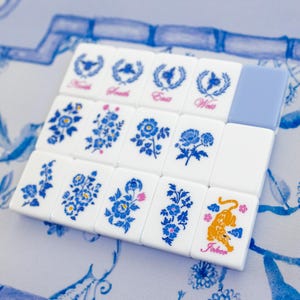 Puede incluir: Un juego de fichas de Mahjong blancas con dise&ntilde;os florales azules y las palabras "Norte", "Sur", "Este" y "Oeste". Una ficha presenta un tigre naranja y la palabra "Joker". Las fichas est&aacute;n dispuestas sobre una superficie con estampado azul y blanco.