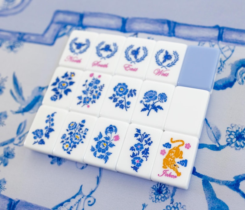 Puede incluir: Un juego de fichas de Mahjong blancas con dise&ntilde;os florales azules y las palabras "Norte", "Sur", "Este" y "Oeste". Una ficha presenta un tigre naranja y la palabra "Joker". Las fichas est&aacute;n dispuestas sobre una superficie con estampado azul y blanco.