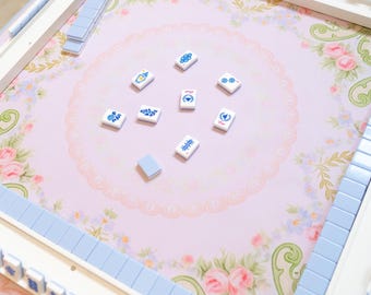 May include: A Mahjong game set with a pink floral patterned playing surface. The set includes white and blue tiles, and white and blue tile racks. The tiles are scattered on the playing surface.