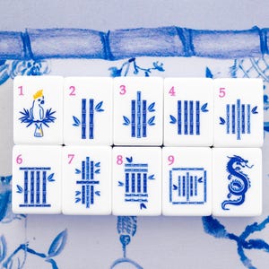 Puede incluir: Un juego de fichas de Mahjong blancas con detalles azules y rosas. Las fichas presentan varias ilustraciones, incluyendo un loro, tallos de bamb&uacute; y un drag&oacute;n. Las fichas est&aacute;n dispuestas sobre una superficie con estampado azul y blanco.