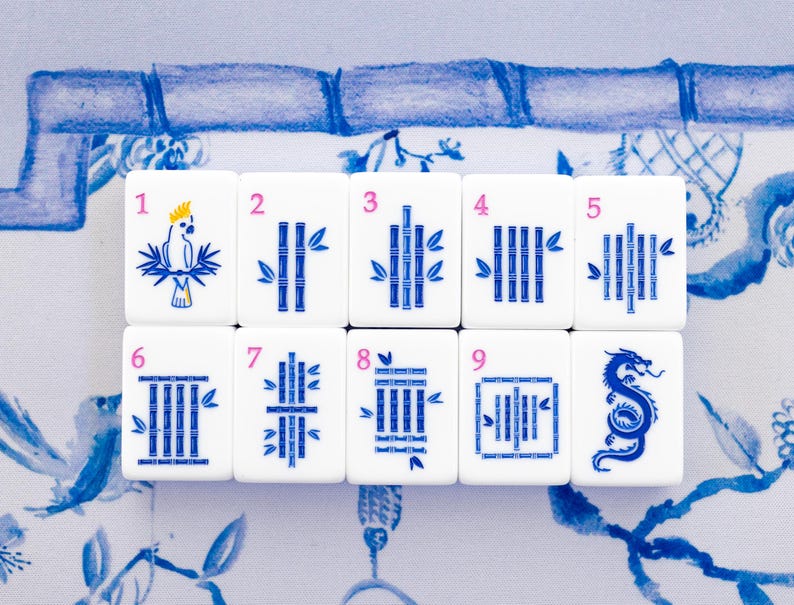 Puede incluir: Un juego de fichas de Mahjong blancas con detalles azules y rosas. Las fichas presentan varias ilustraciones, incluyendo un loro, tallos de bamb&uacute; y un drag&oacute;n. Las fichas est&aacute;n dispuestas sobre una superficie con estampado azul y blanco.