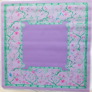 K&ouml;nnte beinhalten: Eine lavendelfarbene Tischdecke mit einem gr&uuml;nen Bambusrand und einem floralen Muster mit rosa Bl&uuml;ten, gr&uuml;nen Bl&auml;ttern und blauen V&ouml;geln. Die Tischdecke ist quadratisch.