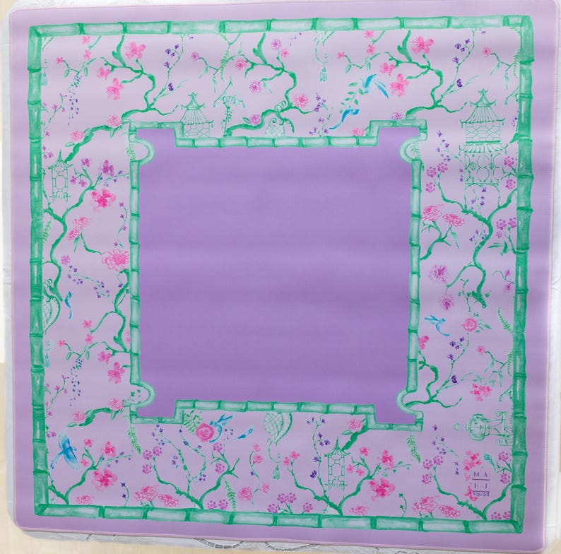 K&ouml;nnte beinhalten: Eine lavendelfarbene Tischdecke mit einem gr&uuml;nen Bambusrand und einem floralen Muster mit rosa Bl&uuml;ten, gr&uuml;nen Bl&auml;ttern und blauen V&ouml;geln. Die Tischdecke ist quadratisch.