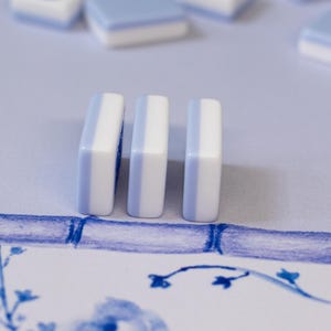 Puede incluir: Tres fichas de juego rectangulares, blancas y azul claro, se alzan sobre una superficie estampada. Hay fichas adicionales dispersas en el fondo. El dise&ntilde;o presenta elementos florales y de estilo bamb&uacute; azules, creando una est&eacute;tica cohesiva.